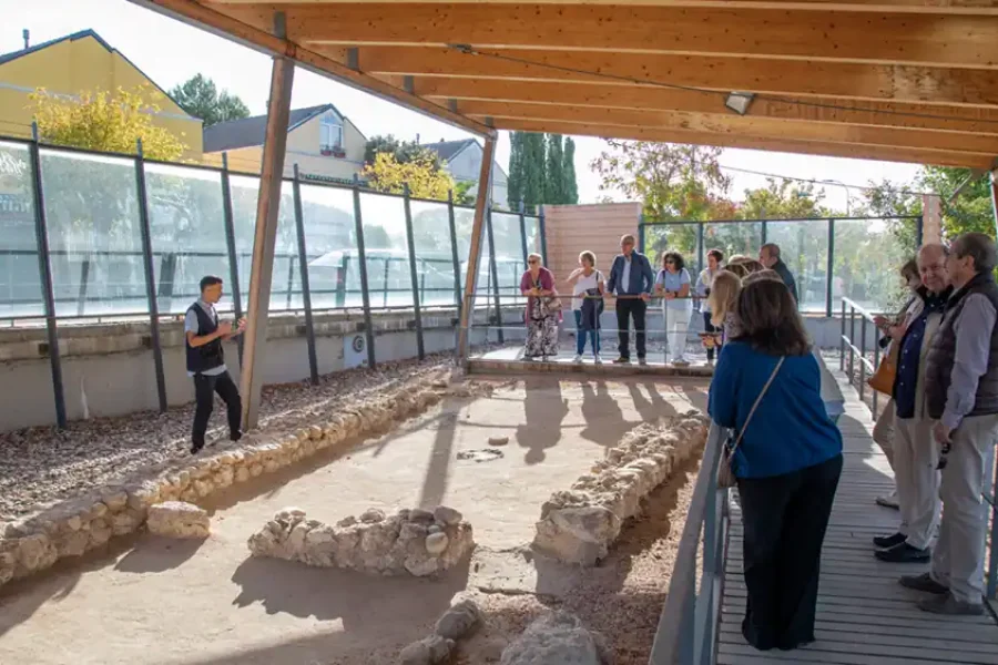Durante una jornada de puertas abiertas, un grupo permanece en una pasarela de madera observando el yacimiento carpetano bajo una estructura exterior cubierta, mientras un guía les habla. La zona cuenta con paneles de cristal y edificios cercanos.