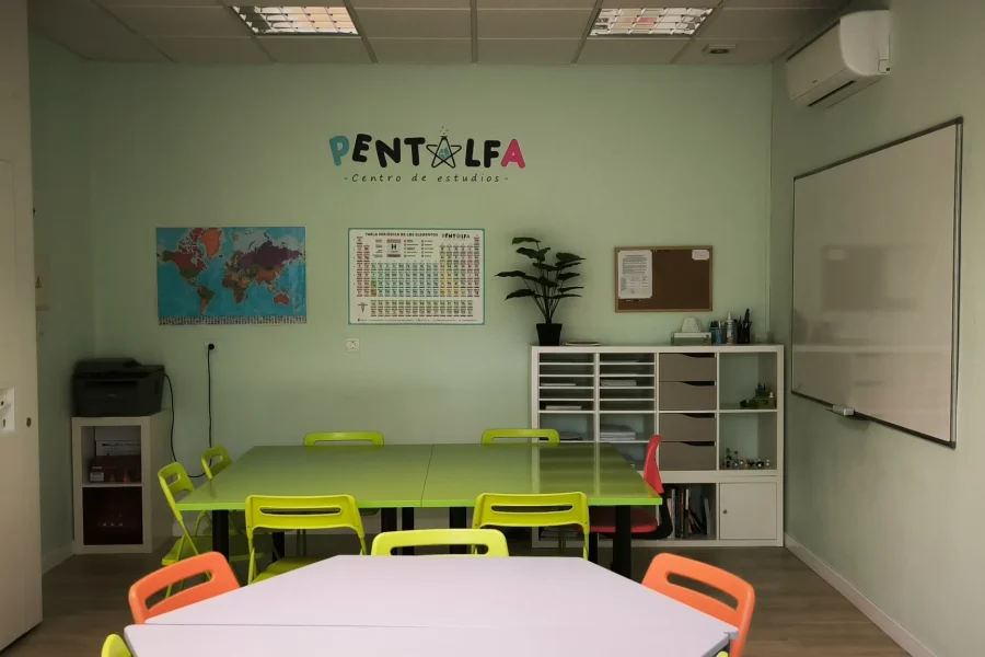Un aula en Rivas con paredes de color verde claro, sillas de colores alrededor de las mesas, un mapamundi, la tabla periódica, una pizarra blanca, estanterías y una pequeña planta. En la pared se lee «Pentalfa Centro de Estudios», ideal para clases de refuerzo en verano.