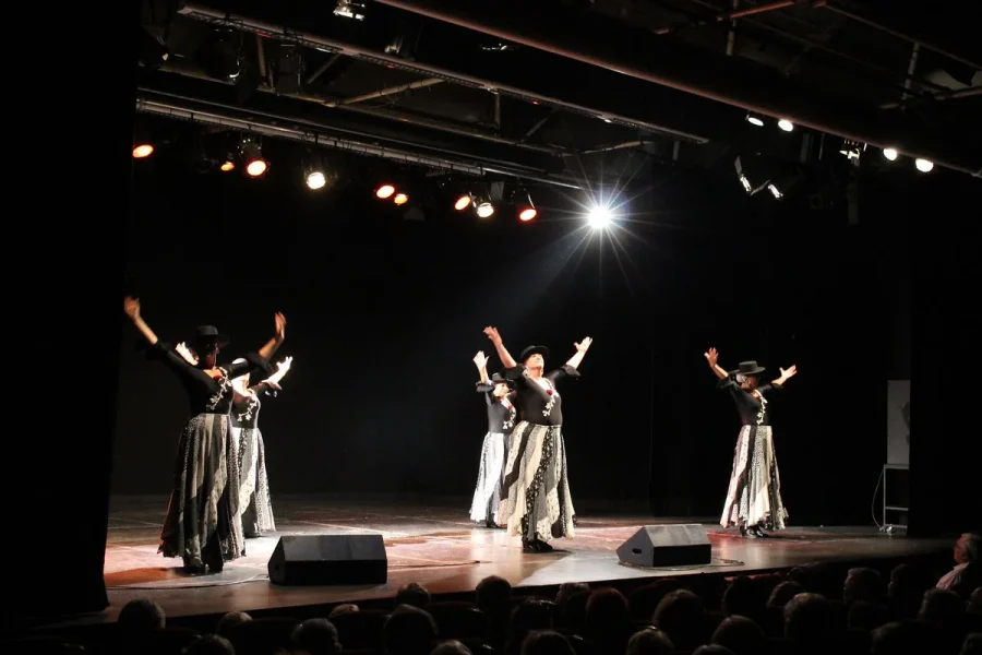 Cinco artistas, con camisetas negras y faldas largas estampadas, bailan en un escenario bajo brillantes focos para promover la solidaridad a través del arte.