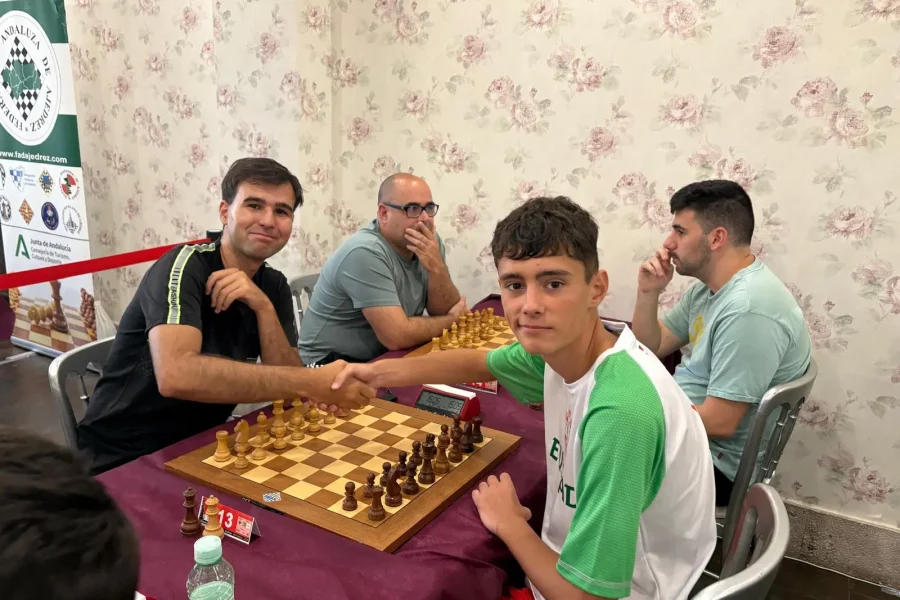 Dos hombres se dan la mano en un tablero de ajedrez al comienzo de una partida del Campeonato de España de ajedrez relámpago, con otros dos sentados en la misma mesa. La escena de interior presenta papel pintado floral de fondo.