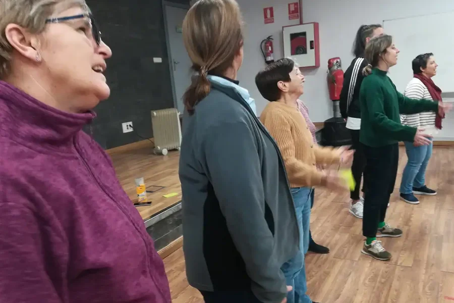 Un grupo de adultos en fila en el interior sobre un suelo de madera, participando en una actividad para talleres. Se ven abrigos y ropa informal, con señales de seguridad contra incendios y equipos en la pared -parte del evento Aula Abierta Mujeres inscritas.