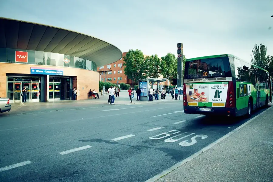 Un autobús urbano operado por el Consorcio Regional de Transportes está aparcado en una parada cercana a la boca de Metro - Urbanizaciones en Rivas. Varias personas pasean o esperan cerca, con árboles y edificios de fondo.