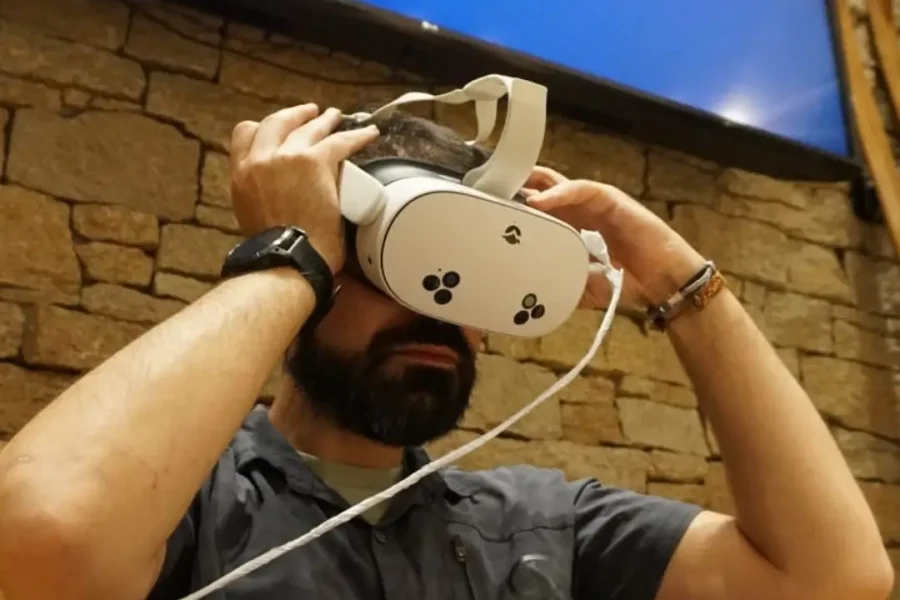 Un hombre con barba lleva unos auriculares de realidad virtual delante de un muro de piedra y una pantalla en la que aparece el logotipo de B4Experience, que explora actividades de aventura. En la parte inferior de la imagen aparecen los logotipos de financiación del Gobierno y de la UE.
