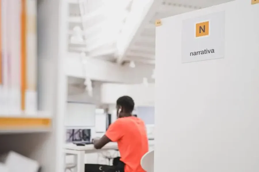 Una persona que lleva una camiseta naranja está sentada ante un escritorio en una luminosa biblioteca de Madrid, trabajando con el ordenador. En primer plano se ven estanterías y un cartel con la inscripción «Narrativa», lo que resalta el animado ambiente de las bibliotecas de Madrid en verano.