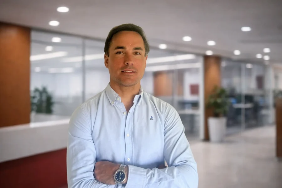 Un hombre de pelo castaño corto con una camisa azul claro abotonada está de pie con los brazos cruzados en una moderna oficina de España con paredes de cristal, plantas en macetas y luces en el techo, que reflejan el espíritu innovador de Fazil Crypto y la normativa MiCA.