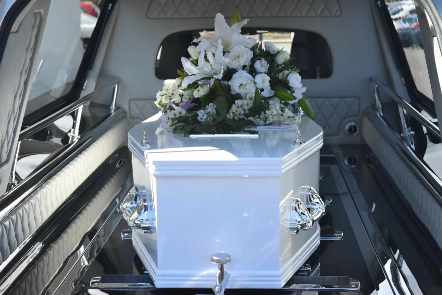 Un ataúd blanco con asas plateadas y un gran arreglo floral en la parte superior se coloca dentro de un coche fúnebre, mientras los asistentes, con aire sombrío, recuerdan en silencio la importancia de conocer técnicas como los primeros auxilios en caso de atragantamiento en momentos de crisis.
