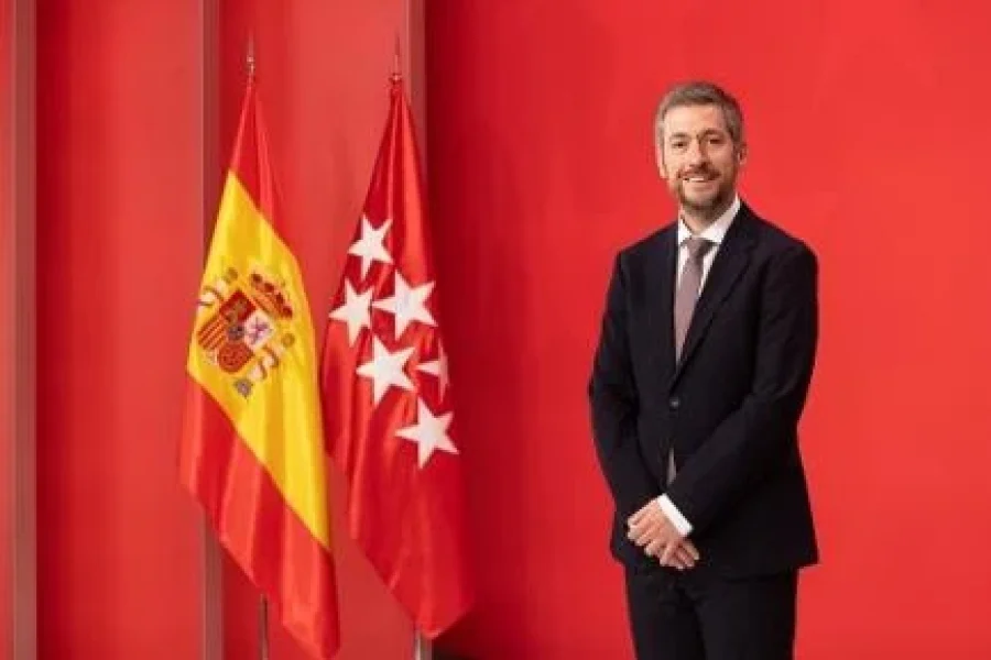 El Consejero Miguel Ángel García, vestido de traje, ante la bandera de España y la bandera de la Comunidad de Madrid, sobre fondo rojo.