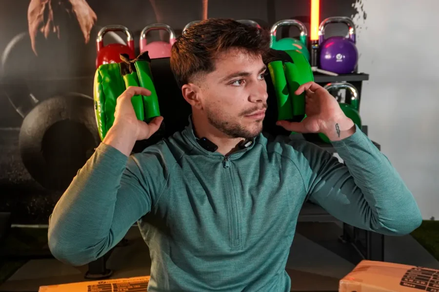 Un hombre con una sudadera verde con capucha sostiene dos pesas verdes a la altura de los hombros en un gimnasio; al fondo se ven pesas rusas y material de gimnasio.