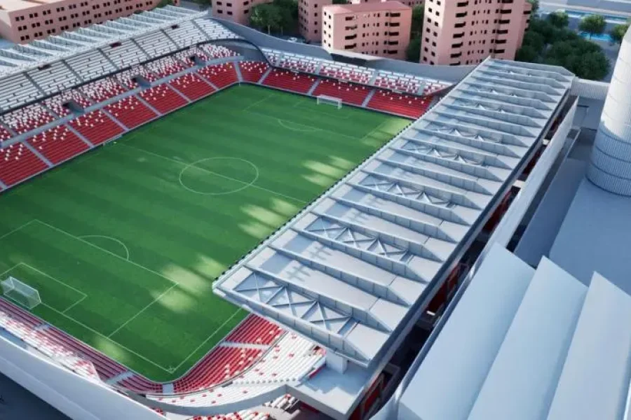 Vista aérea del Estadio de Vallecas, en la que se aprecian los asientos rojos y blancos, el campo verde, las gradas parcialmente cubiertas, los edificios altos cercanos y el logotipo de la Comunidad de Madrid en la esquina superior derecha, en medio de las obras de reforma en curso.