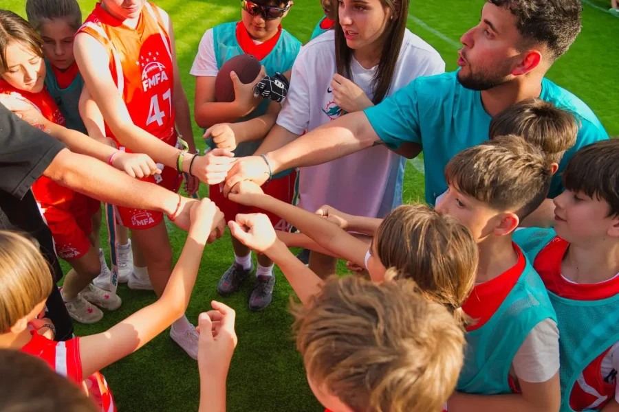Un grupo de niños y dos adultos están de pie formando un círculo sobre la hierba, estirando los brazos hacia el centro para chocar los puños entre ellos. Los niños llevan petos deportivos y hace sol, lo que crea una escena perfecta para la diversión de un campamento de verano inspirado en los Miami Dolphins.