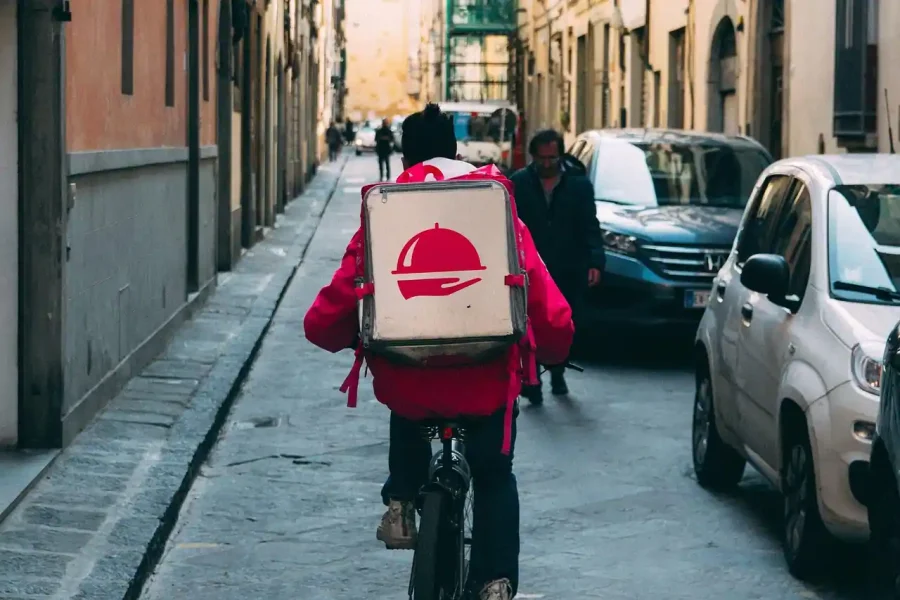 Un repartidor de comida a domicilio vestido con una chaqueta roja recorre en bicicleta una estrecha calle de la ciudad con coches aparcados y peatones, portando una gran bolsa aislante de reparto con el logotipo de un servicio de comida a domicilio, entregando comida sana a personas mayores en situación de vulnerabilidad.