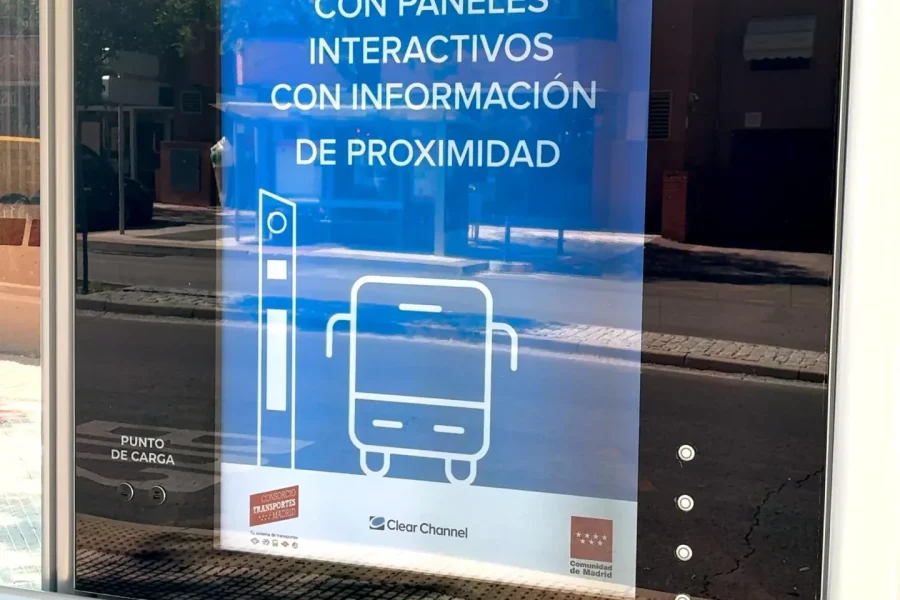 Un cartel en una parada de autobús, redactado en español, anuncia 10 marquesinas inteligentes con paneles interactivos que ofrecen información de proximidad e incluyen mapas interactivos. El cartel incluye iconos de un autobús y un punto de información, mientras que se aprecian los edificios cercanos y la acera de Rivas.