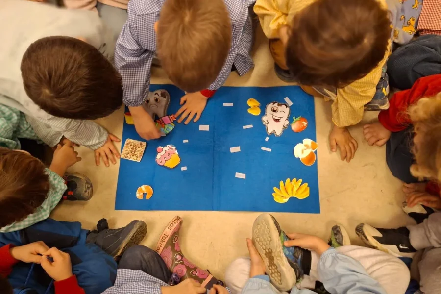 Un grupo de niños está sentado en círculo en el suelo, alrededor de una pizarra azul con dibujos animados y tarjetas, concentrados en una actividad que realizan juntos, lo que refleja el sentido de comunidad que a menudo se fomenta en las escuelas infantiles de Rivas.