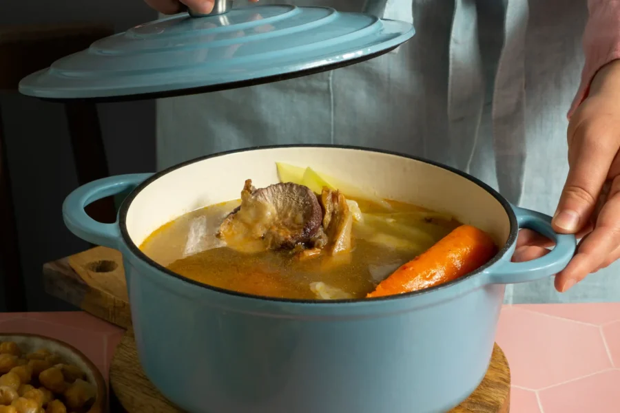 Una persona con un delantal azul claro levanta la tapa de una olla azul con sopa, dejando ver carne, una zanahoria y apio. Inspirada en la Ruta del Cocido 2026, la escena evoca sabores tradicionales celebrados en restaurantes de toda España.