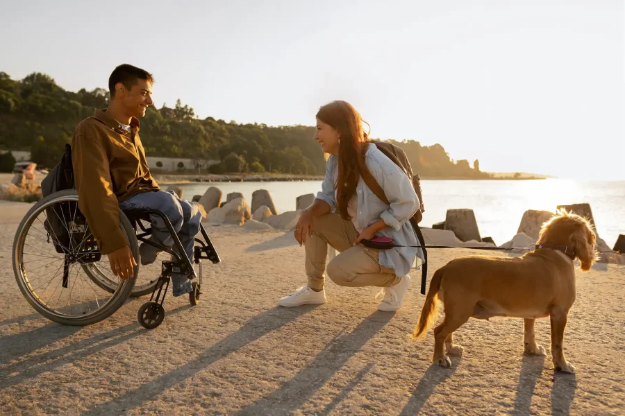 Un hombre con movilidad reducida en silla de ruedas y una mujer arrodillada junto a un perro marrón con correa están al aire libre cerca del agua al atardecer, hablando y sonriendo. Al fondo se ven grandes rocas y la luz del sol.