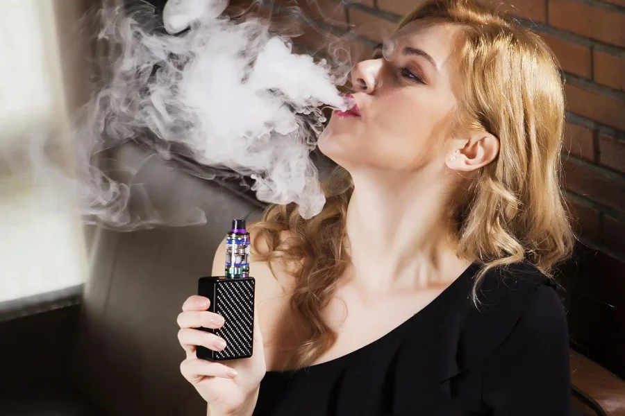 Una mujer de pelo rubio, sentada en un interior, sostiene un cigarro electrónico negro y exhala una gran nube de vapor. Lleva un top negro y está situada cerca de una ventana y una pared de ladrillo.