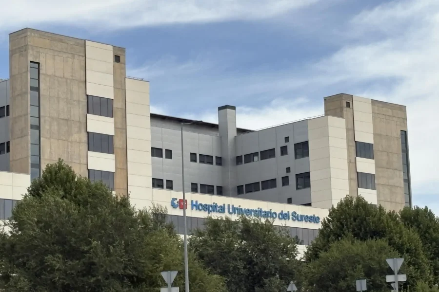 Un edificio hospitalario grande y moderno, con fachadas de color beige y blanco, numerosas ventanas y un letrero en el que se lee «Hospital Universitario del Sureste» —visible en parte tras los árboles verdes bajo un cielo parcialmente nublado—, parece utilizarse tanto para la atención a los pacientes como para tareas administrativas, como el «borrador automático».
