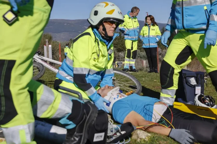 Efectivos de emergencias médicas del SUMMA 112 atienden a un ciclista herido que yace en el suelo con un collarín en la Comunidad de Madrid, rodeado de equipos médicos y otros efectivos con uniformes brillantes.