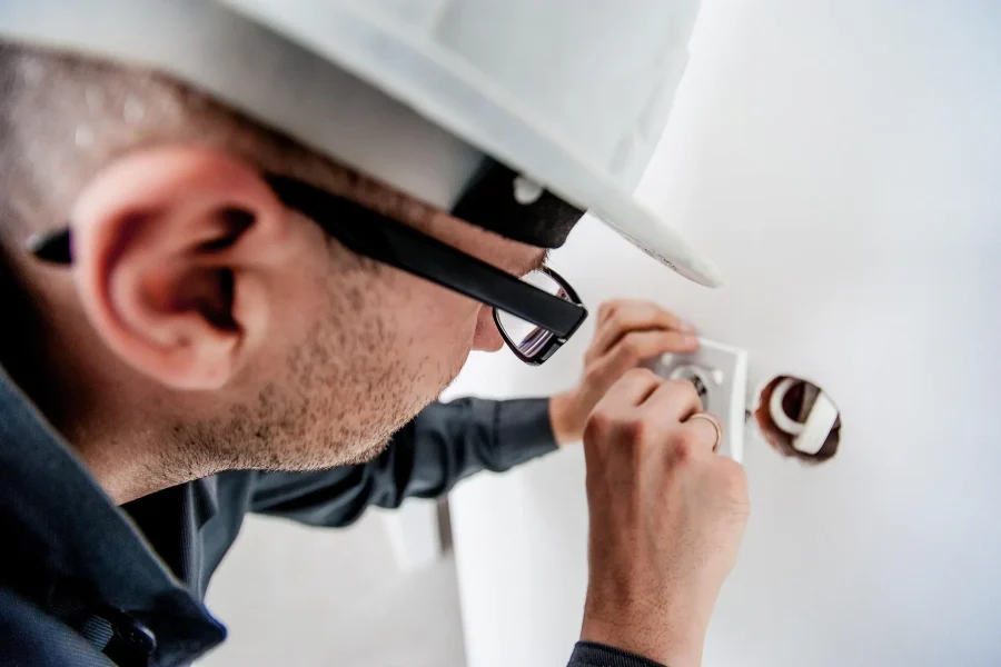 Un electricista, con casco y gafas, realiza tareas de mantenimiento en la instalación eléctrica de una vivienda mientras instala una toma de corriente en la pared.