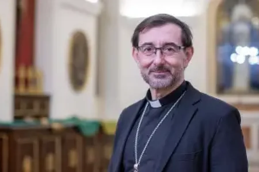 Un hombre con gafas y alzacuello clerical se encuentra en el interior, vestido con un traje negro y un collar con una cruz, tal vez parecido al arzobispo de Madrid. El fondo revela la decoración religiosa y los detalles borrosos de la convivencia eclesiástica.