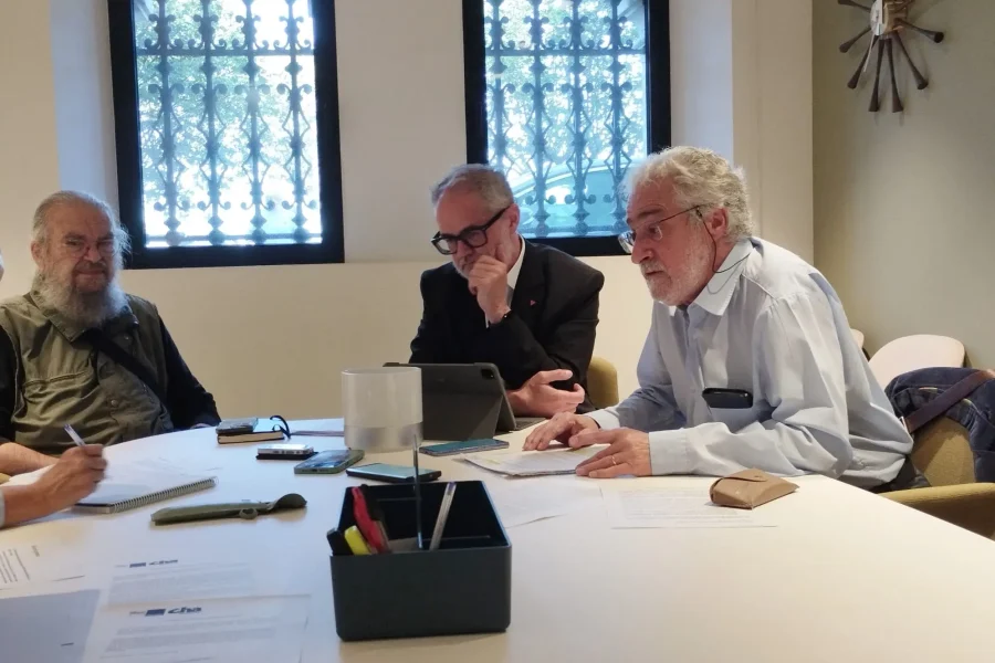 Tres hombres mayores se sientan a una mesa de conferencias en una sala con dos ventanas enrejadas, discutiendo inmatriculaciones que afectan a la Iglesia Católica. Sobre la mesa hay papeles, bolígrafos y una tableta mientras uno escribe, otro comprueba un dispositivo y el tercero mira a la cámara.