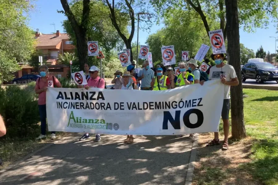 Un grupo de personas de pie en una acera de Rivas, sosteniendo carteles con el símbolo "no" y una pancarta en la que se lee "Alianza Incineradora de Valdemingómez NO", exigiendo el cierre de Valdemingómez. Al fondo aparecen árboles y casas.