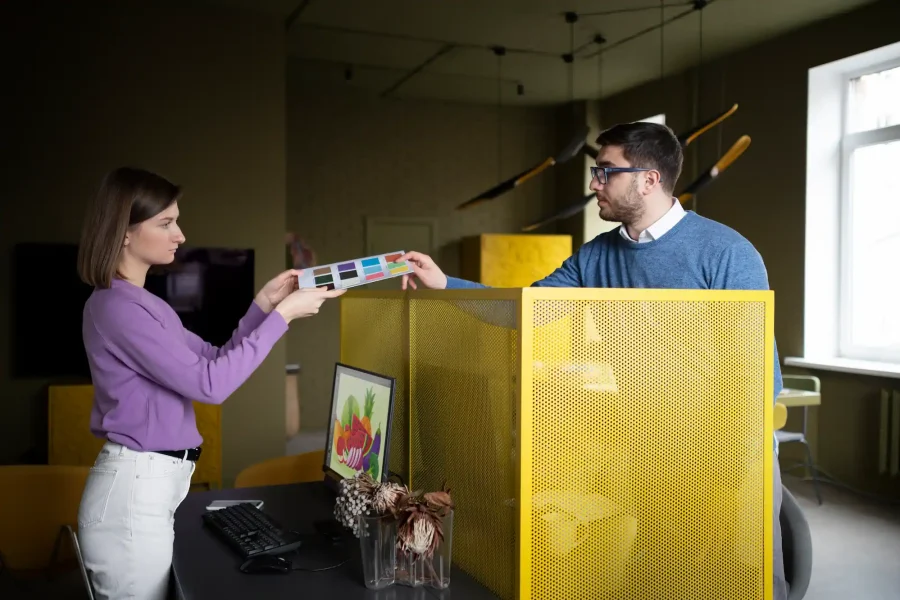 Dos personas en una oficina, probablemente de una agencia de marketing, examinan juntas una muestra de color; una mujer sujeta un extremo y un hombre se sienta en un escritorio sujetando el otro, con un ordenador y un separador amarillo entre ellos.