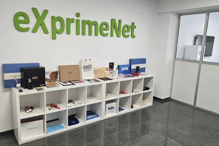 Una estantería blanca con diversos artículos promocionales y embalajes se despliega contra una pared con el logotipo verde de eXprimeNet, destacando nuestra consultora digital. A la derecha hay una puerta de cristal y una ventana, y debajo un suelo oscuro y brillante.