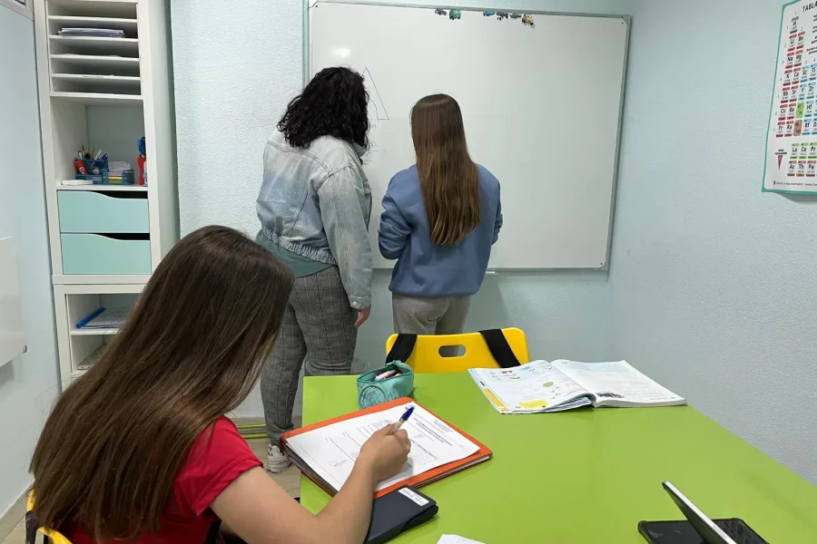 Tres chicas están en un aula de la Academia Pentalfa de Rivas. Dos están de pie junto a una pizarra, mientras que una está sentada en una mesa verde escribiendo en un cuaderno, inmersa en un Intensivo de PAU. Sobre la mesa hay libros abiertos, un estuche y un teléfono.