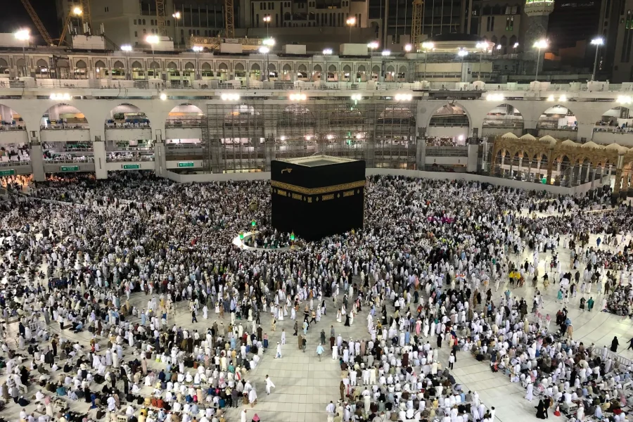 Miles de personas se reúnen en torno a la Kaaba de la Gran Mezquita de La Meca, vestidas en su mayoría de blanco, para peregrinar a La Meca. La mezquita está brillantemente iluminada y rodeada de arcos de varios pisos. Crece la preocupación por el golpe de calor debido al cambio climático.
