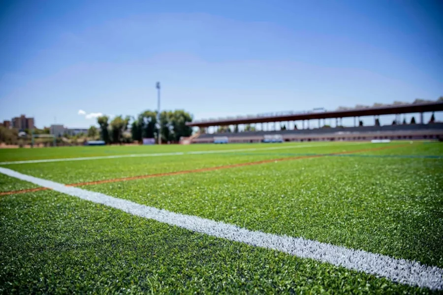 Una imagen en primer plano de un campo deportivo de césped artificial verde, mantenido por una máquina de corte automática, muestra líneas blancas y rojas. A lo lejos se ven árboles, un edificio y una farola, bajo un cielo azul claro.