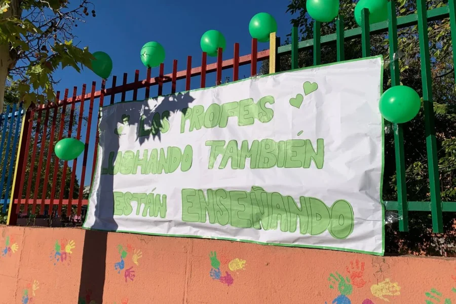 En una valla hay una pancarta en la que se lee Los profes luchando también están enseñando, rodeada de globos verdes que representan la marea verde y de coloridas huellas de manos en la pared que hay debajo.