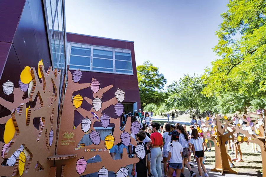 Un grupo de personas, entre las que hay niños, se reúne frente a un edificio moderno, cerca de unos árboles de cartón decorados con bellotas de papel de colores, en uno de los colegios de Rivas que participan en el proyecto 50/50 de ahorro de energía. La escena, luminosa y soleada, transmite un ambiente de comunidad y expectación ante el curso 2024/2025.