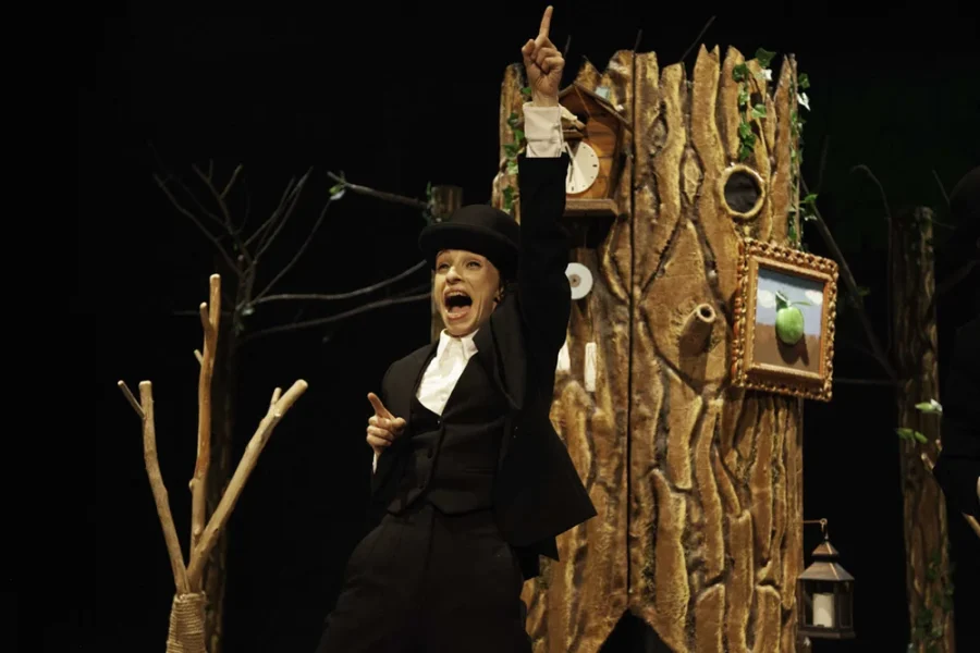 Un intérprete, vestido con traje y sombrero hongo, hace un gesto hacia arriba durante una obra de danza-teatro, frente a un árbol de atrezo al que están colgados un reloj y una fotografía enmarcada de una manzana.