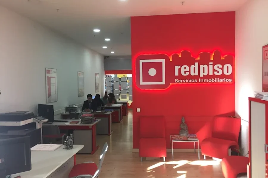 Una moderna agencia inmobiliaria con una decoración en tonos rojos y blancos. El espacio está decorado con sillas y escritorios rojos. El logotipo de «redpiso Servicios Inmobiliarios» aparece en una pared destacada de color rojo brillante. En el fondo, varias personas trabajan en sus escritorios.