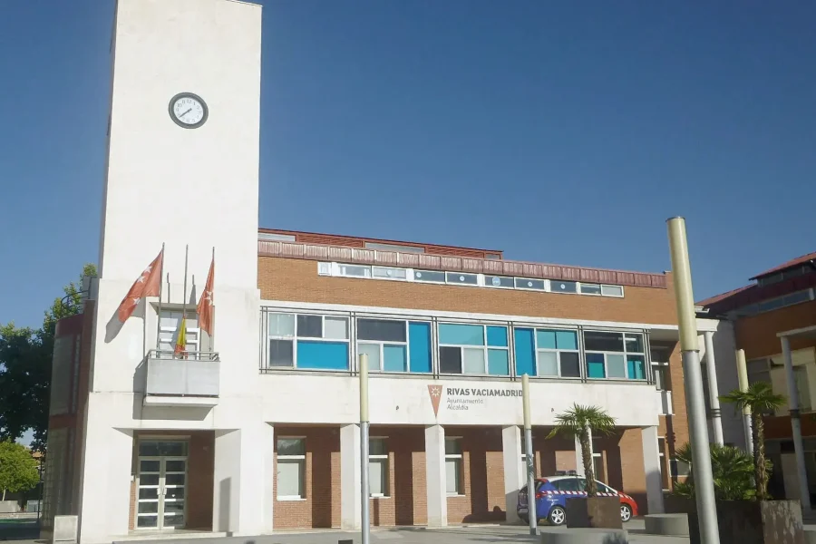 Un edificio de dos plantas del Ayuntamiento de Rivas con una torre del reloj, varias banderas y un cartel en el que se lee Rivas Vaciamadrid Alcaldía; un coche de policía está aparcado fuera bajo un cielo azul despejado.