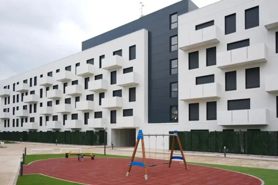 Un moderno edificio de apartamentos blanco con contraventanas negras, construido en el marco del Plan Estatal de Vivienda, se alza detrás de un pequeño parque infantil con columpios y juegos elásticos sobre un suelo de caucho rojo. El cielo está nublado.