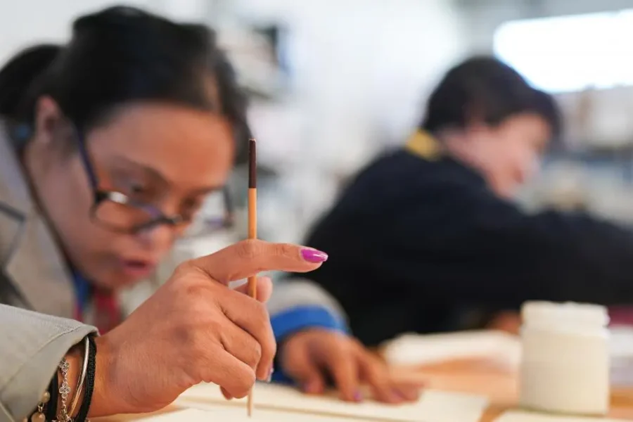 Una persona con un pincel parece estar pintando sobre papel, mientras otra trabaja en una mesa de un aula o estudio de arte. Los materiales artísticos son visibles, lo que refleja cómo los talleres creativos pueden ser beneficiosos para las personas con discapacidad en la Comunidad de Madrid.