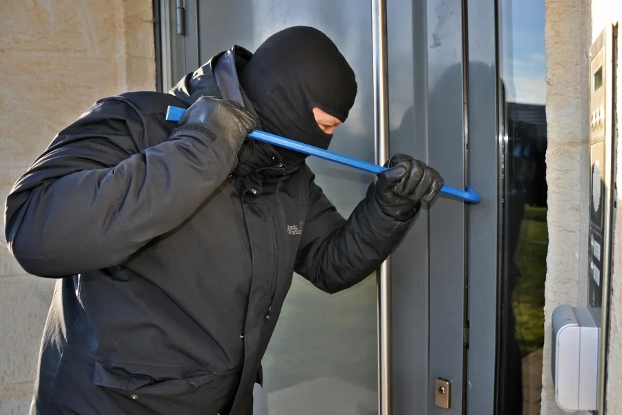 Una persona que lleva una máscara negra, guantes y una chaqueta utiliza una palanca azul para forzar una puerta de cristal, lo que pone de relieve la importancia de reforzar la seguridad para proteger su vivienda en el este de Madrid.