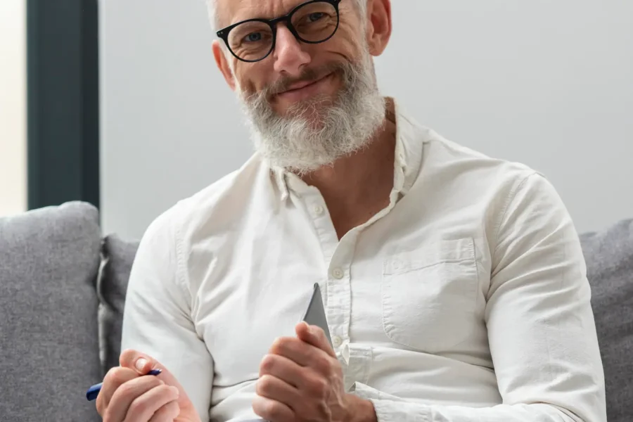 Un hombre de pelo y barba canosos, con gafas y camisa blanca de botones, sentado en un sofá gris con un cuaderno y un bolígrafo en la mano, reflexiona sobre la jubilación anticipada para los trabajadores con más de 40 años cotizados y mira a la cámara con una leve sonrisa.