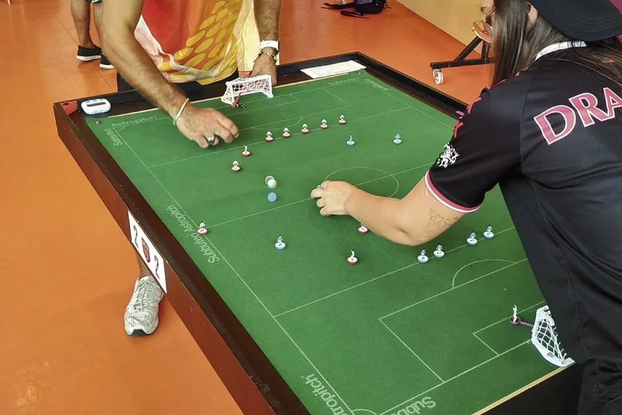 Dos personas juegan a un juego de fútbol de mesa llamado Subbuteo en un tablero de campo verde. El jugador de la izquierda lleva una camiseta naranja, mientras que el de la derecha lleva una camiseta y una gorra negras. En el campo se colocan pequeñas figuritas.