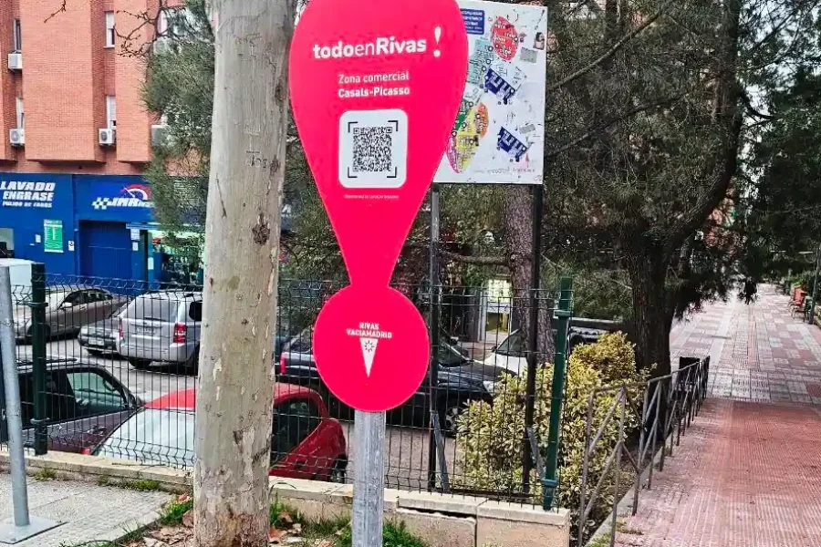 Un cartel rojo en forma de signo de exclamación con un código QR y el texto todoenRivas se encuentra en una acera cerca de coches aparcados, un árbol y un edificio de ladrillo en una zona urbana.
