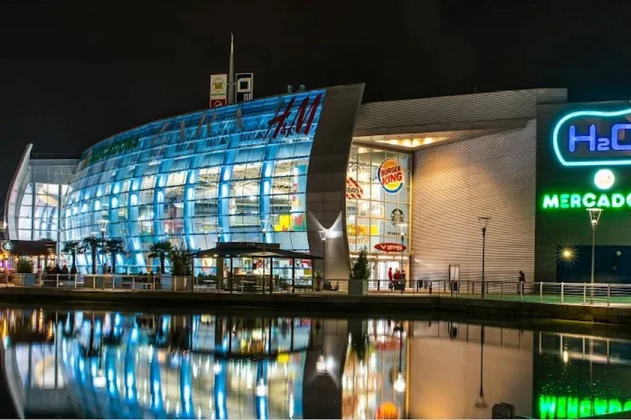 Un moderno centro comercial de Rivas por la noche, con paredes acristaladas, luces de colores y letreros de tiendas como el centro comercial H2O y Mercadona, reflejados en una tranquila superficie de agua en primer plano. El ambiente animado se ve realzado por el resplandor de una pantalla final del Mundial que ilumina la escena.