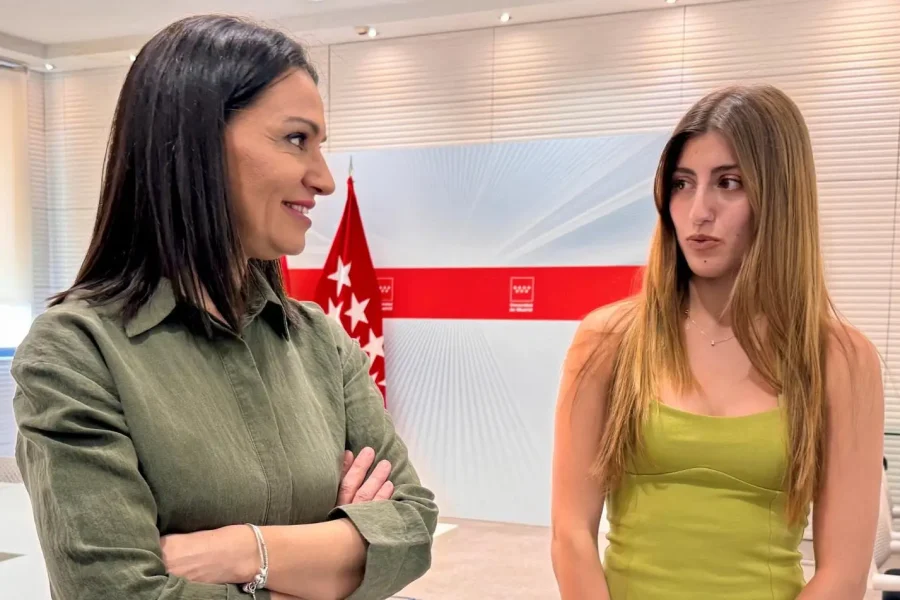 Dos mujeres están de pie en el interior de una habitación, conversando. Una de ellas, Valeria Fragola, de pelo oscuro y camiseta verde, tiene los brazos cruzados; la otra tiene el pelo largo castaño claro y lleva un vestido sin mangas de color verde claro. Al fondo se ve una bandera de la PAU de Madrid.