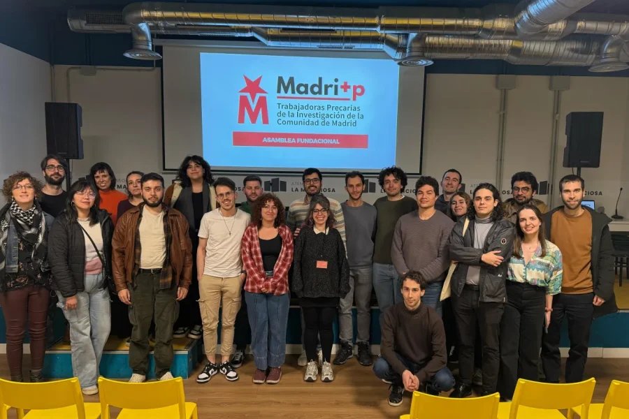 Un grupo de unas 24 personas posa para una foto de grupo delante de una pantalla en la que se ve un logotipo rojo y blanco y un texto en español sobre la asamblea de investigadores de Madrid, en la que se debate el tema del borrador automático. En primer plano se ven sillas amarillas.