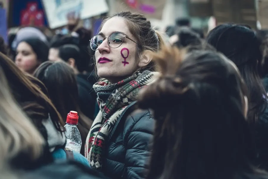 Mujeres que transforman. Una mujer con gafas y pañuelo sujeta una botella entre la multitud. Lleva un símbolo feminista morado dibujado en la mejilla, una poderosa imagen captada por Borrador automático durante una reunión pública o una protesta.